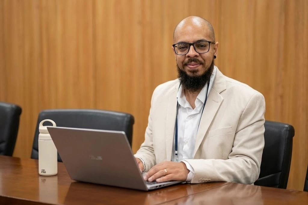 Danilo Souza sentado à mesa diante de um notebook, em uma sala com painéis de madeira.
