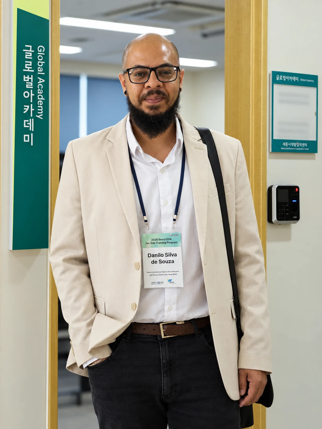 Danilo Souza em pé, usando óculos e blazer claro, diante da entrada da Global Academy durante um programa internacional em Seul.